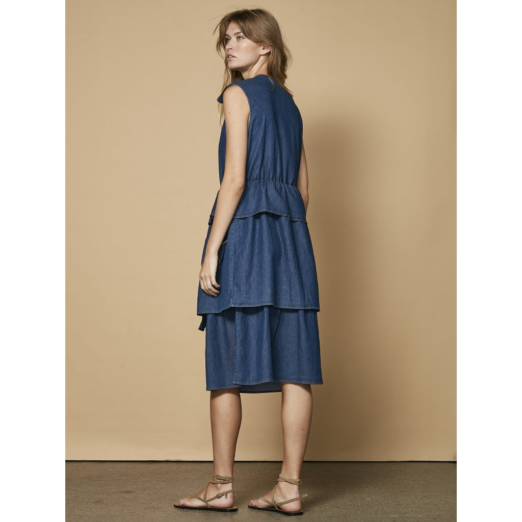 ryggen af model med ærmeløs blå denimkjole med skørt i flere lag. NÜ Ziga denimkjole