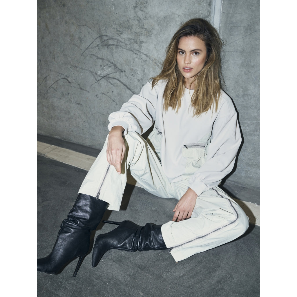 model siddende med langærmet hvid bluse med ribbet struktur, lynlåslommer foran og en let markeret talje. NÜ Zinne Blouse Bone